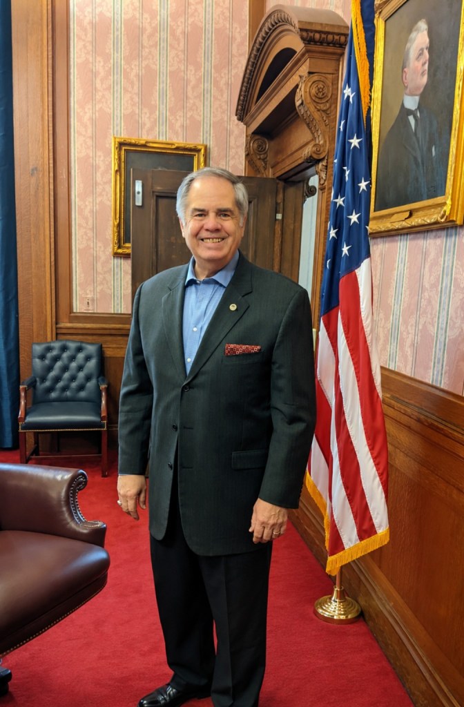 Martin L. Marty Flask standing beside an American flag in a formal municipal office.